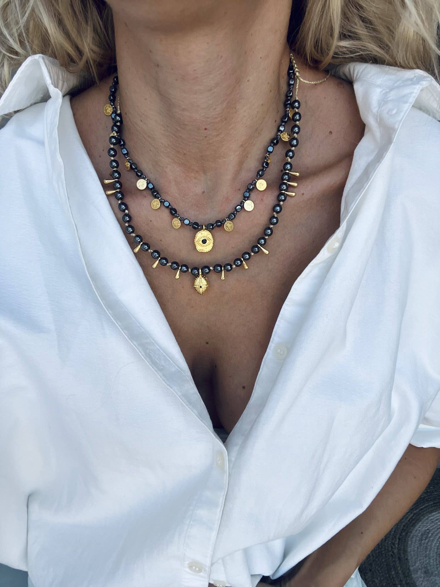 Black hematite necklaces with gold charms layered over a white shirt and jeans.

